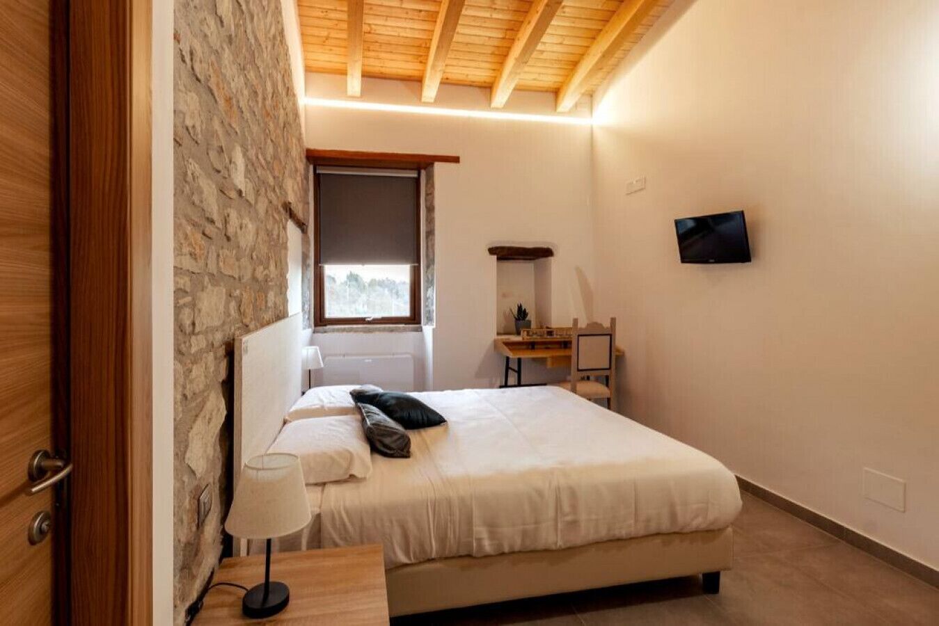 A bedroom with a stone wall, a bed with white bedding, a bedside table with a lamp, a small desk with a chair, and a TV mounted on the wall.