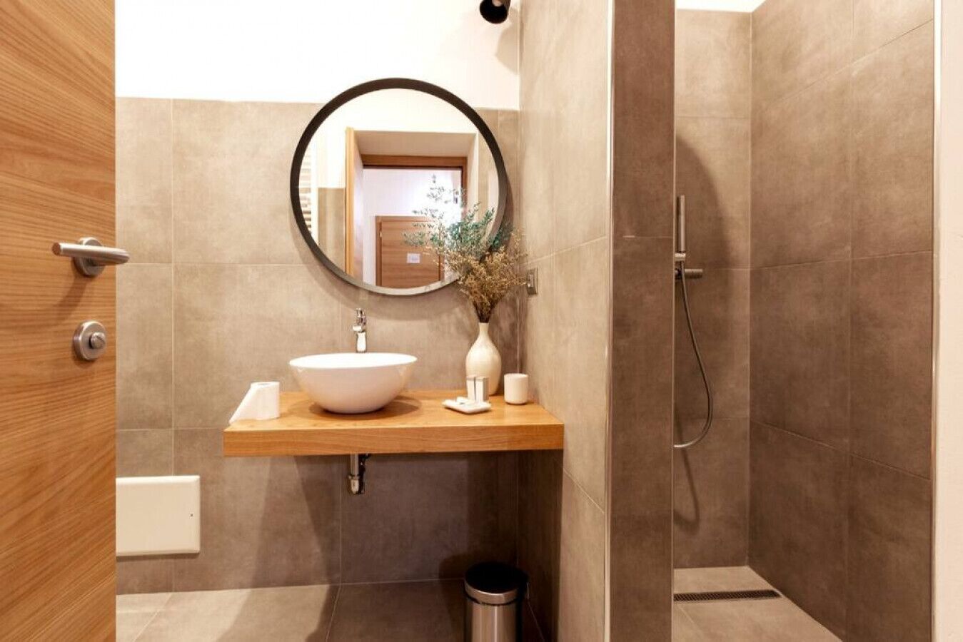 A modern bathroom with a wooden vanity, a round mirror, a white sink, and a shower area.