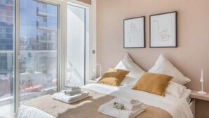 A modern bedroom with a large window, a bed with white linens, two framed artworks on the wall, and a bedside table with a lamp. - Cozy 1BR with balcony sea view (Dubai)