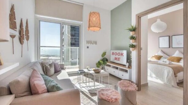 A modern living room with a sofa, coffee table, and a TV. There is a balcony with a view, and a bed with a 'BOOK ME' sign. - Cozy 1BR with balcony sea view (Dubai)