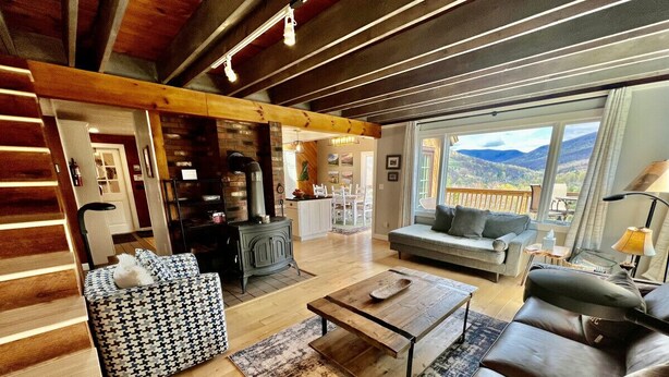 Un salón amplio con suelo de madera, un sofá, una mesa de centro y vistas a las montañas a través de grandes ventanales.
