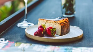 A slice of cake with strawberries on a plate, a glass of white wine, and a potted plant on a table. - K-Log Studios  (Kas)