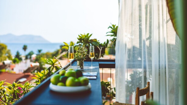 A balcony with a table set for two, featuring a plate of green apples and two glasses of white wine, overlooking a scenic view of the sea and mountains. - K-Log Studios  (Kas)