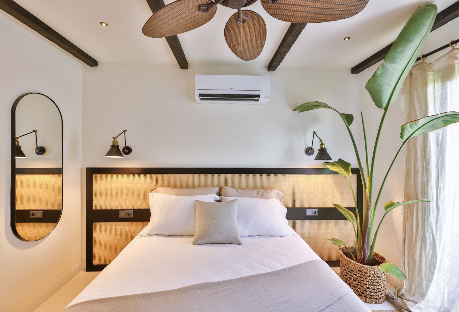 Ein Hotelzimmer mit einem großen Bett, einem Deckenventilator, einer Klimaanlage, einem Spiegel, zwei Wandlampen und einer Zimmerpflanze.