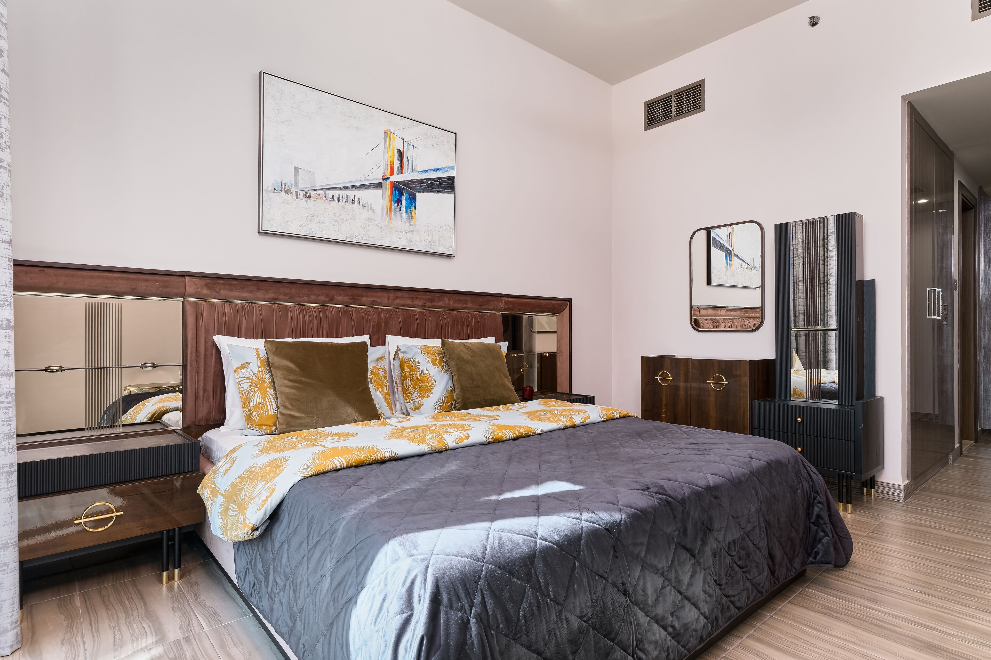 Ein modernes Hotelzimmer mit einem großen Bett, einer Kopfteil mit Spiegel, einem Schminktisch und einem Gemälde an der Wand.