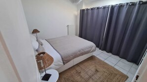 Ein Schlafzimmer mit einem Bett, einem kleinen Tisch, einer Lampe, einem Fenster mit Vorhängen und einem Oberlicht.