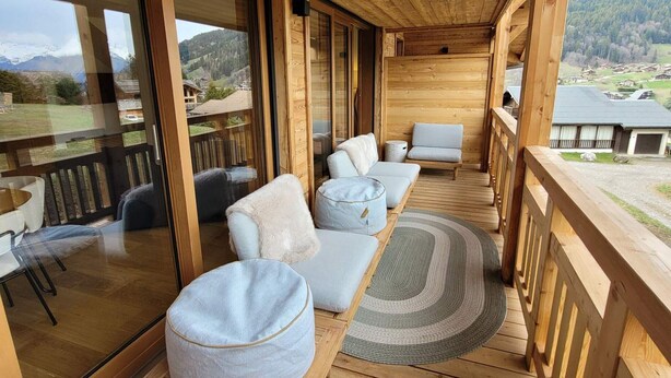 Una terraza de madera con un sofá, un reposapiés, ventanas amplias y vistas a un paisaje montañoso.