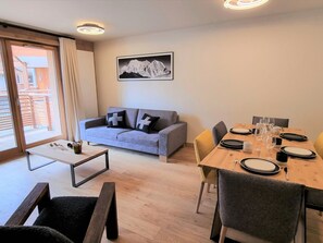 Un salon moderne avec un canapé gris, une table à manger dressée pour quatre personnes et une grande fenêtre avec des rideaux.