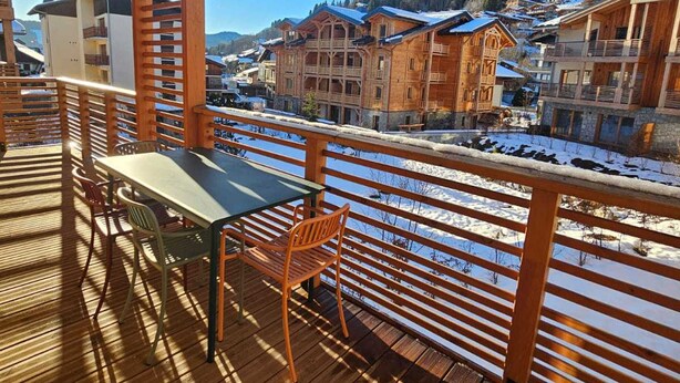 A wooden deck with a table and chairs, overlooking a snowy village with wooden buildings.