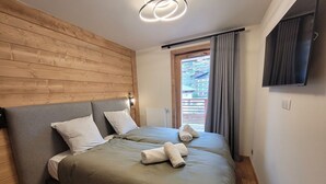 A bedroom with a wooden headboard, a bed with grey bedding, a flat-screen TV on the wall, and a window with curtains.