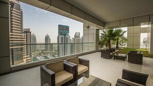 A modern balcony with wicker furniture, a view of high-rise buildings, and a glass railing. - Beautiful 1BR with sunset view (Dubai)