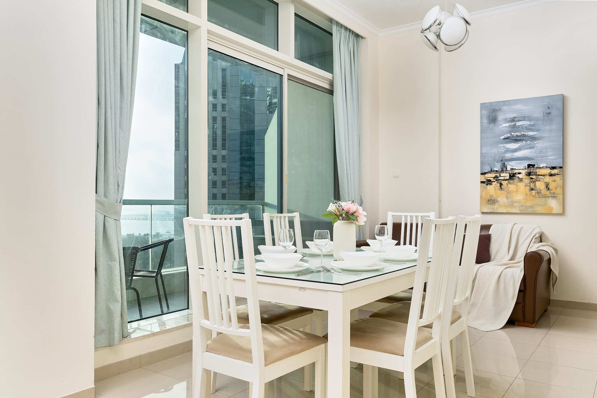 A dining area with a glass table, white chairs, and a view of buildings through large windows.