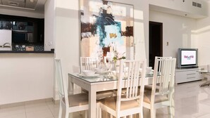 A modern dining area with a glass table and white chairs, a kitchen with dark cabinets, and a TV displaying "Welcome Home". - Beautiful 1BR with sunset view (Dubai)