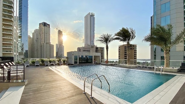 A rooftop pool with a wooden deck, surrounded by high-rise buildings and palm trees. - Beautiful 1BR with sunset view (Dubai)