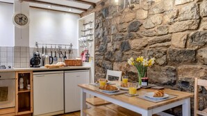 A cozy kitchen with a stone wall, wooden beams, and a breakfast table set with croissants, juice, and a vase of flowers.
