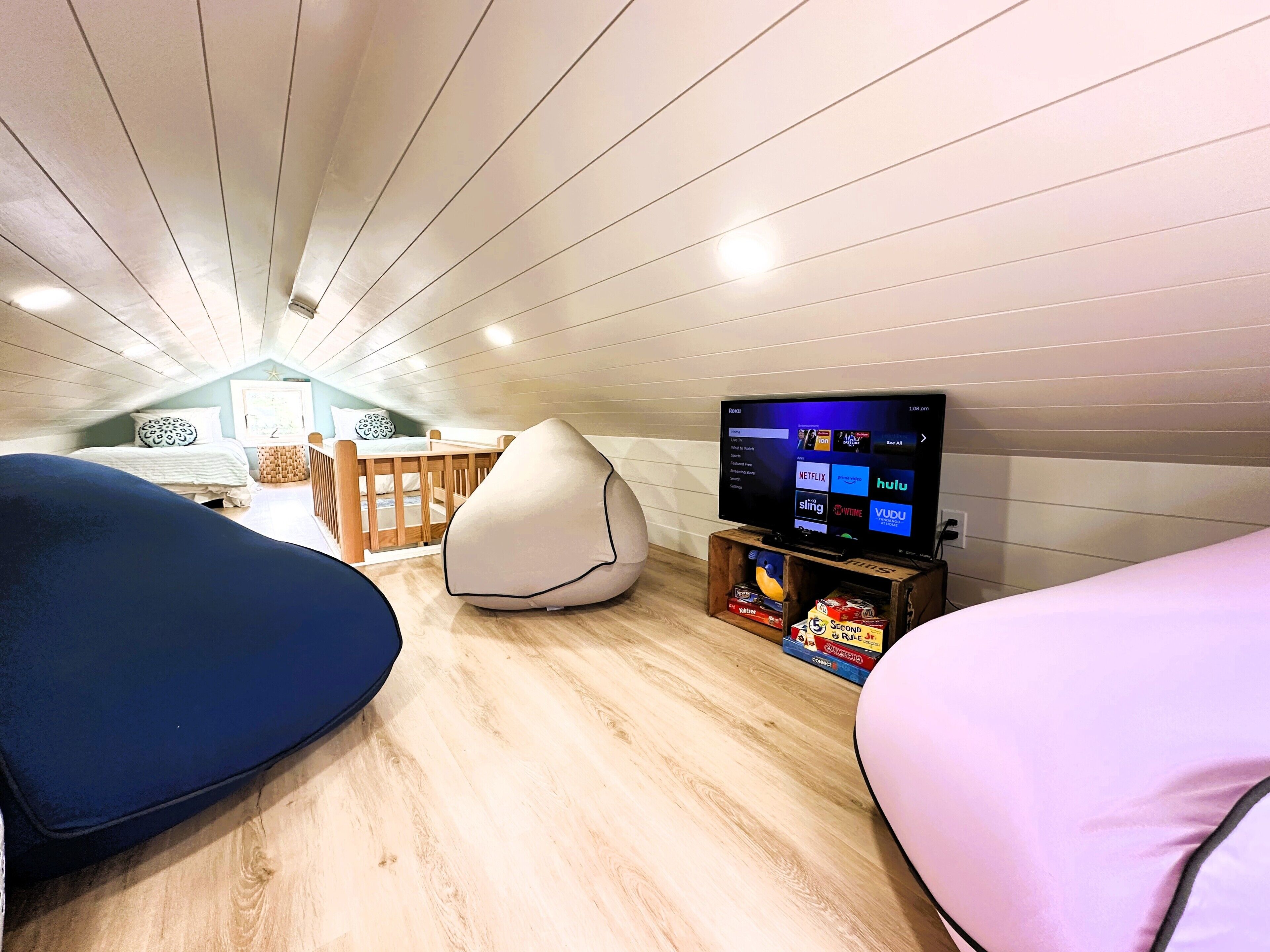 Een knus zolderkamer met een schuine kap, een houten bed, een relaxfauteuil, een kleine televisie op een houten dressoir, en een boekenkast met boeken en spellen.