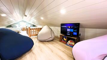 Een knus zolderkamer met een schuine kap, een houten bed, een relaxfauteuil, een kleine televisie op een houten dressoir, en een boekenkast met boeken en spellen.