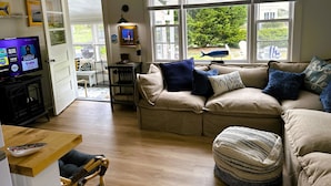 Un salón con un sofá color beige, una mesa de centro de madera y un televisor sobre un mueble bajo. Se observan ventanas con persianas, una lámpara de techo y una mesita con lámpara.