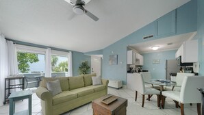 A living room with a green sofa, a wooden chest, and a dining area with white chairs and a glass table.