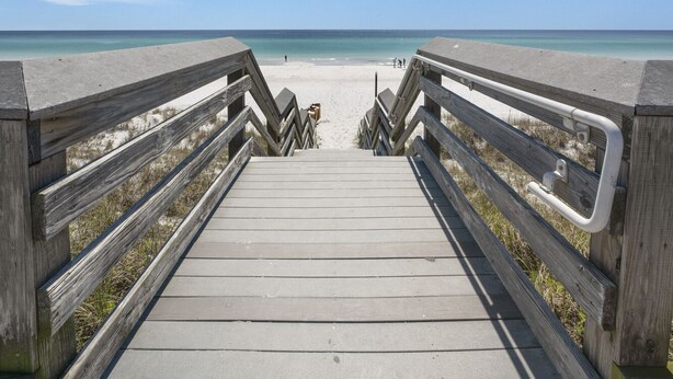A wooden boardwalk leading to a beach with a clear blue sky and scattered clouds.