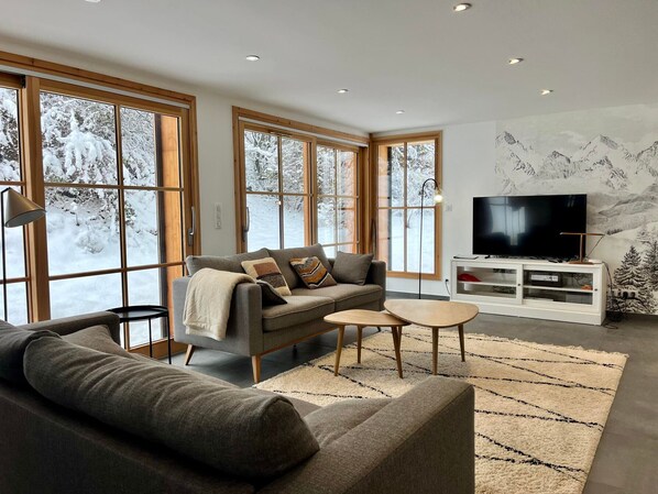 A modern living room with a grey sofa, a coffee table, and a TV on a white cabinet. Large windows with wooden frames offer a view of snow-covered trees.