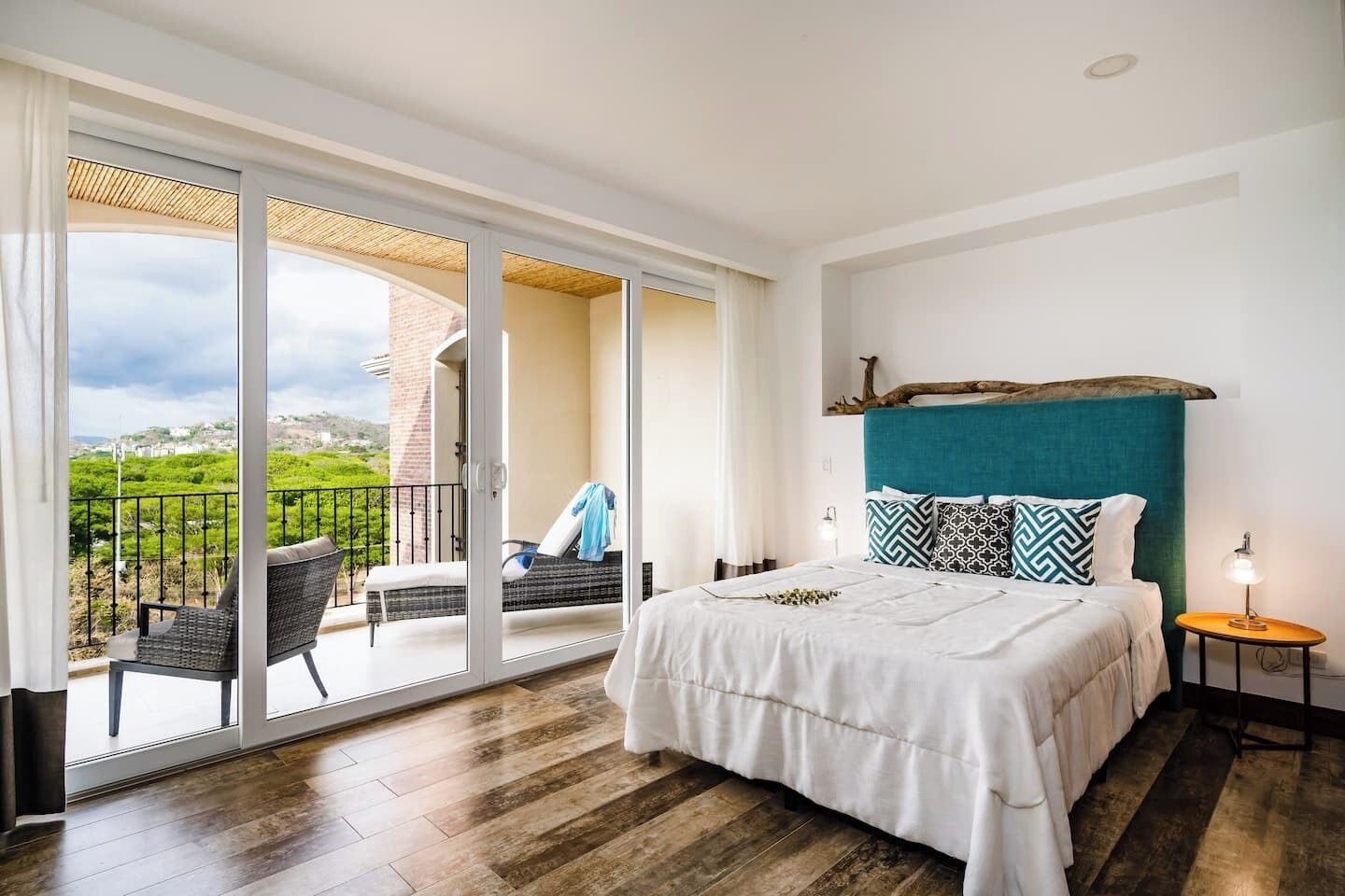 Une chambre avec une grande porte-fenêtre coulissante donnant sur un balcon, un lit avec une tête de lit de couleur turquoise et une petite table de chevet avec une lampe.
