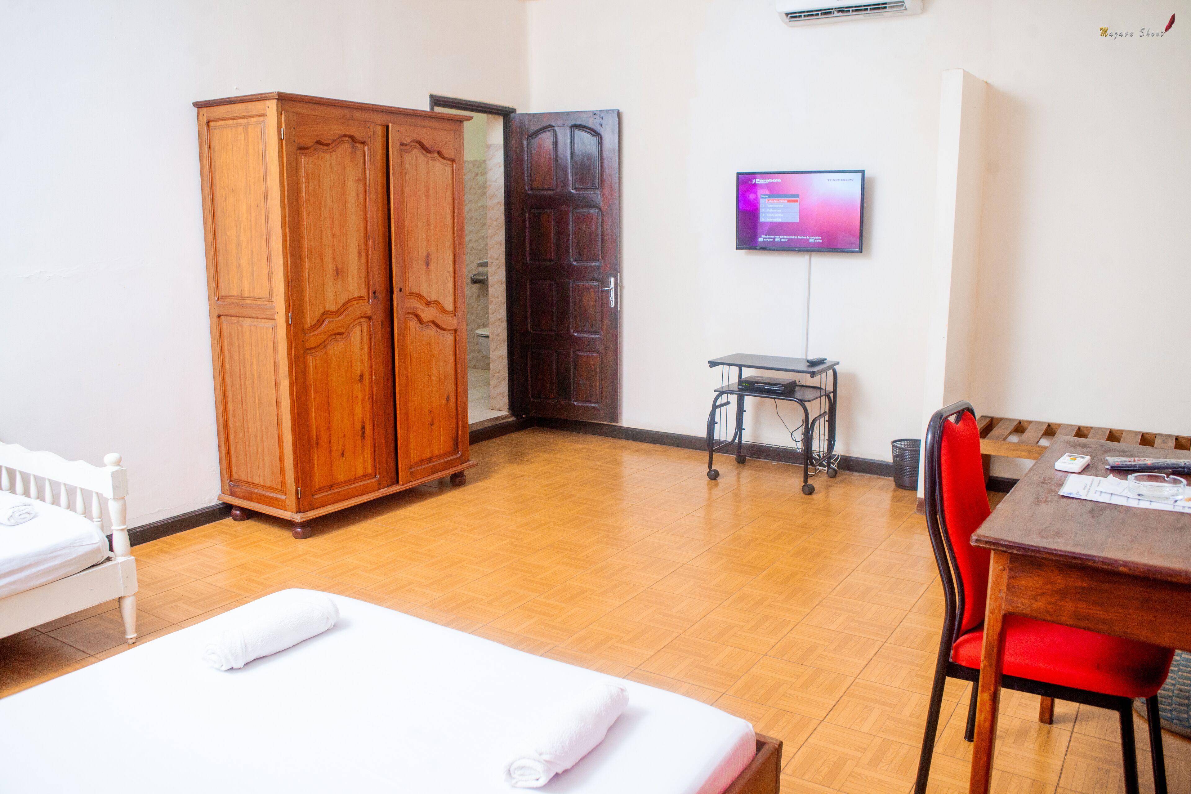 A hotel room with a bed, a wooden wardrobe, a desk with a chair, and a television on the wall.