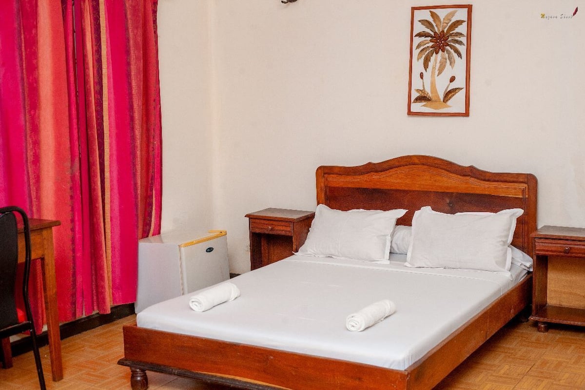 Una cama bien hecha con sábanas y almohadones blancos, un cabecero de madera y una mesita de noche con una lámpara.