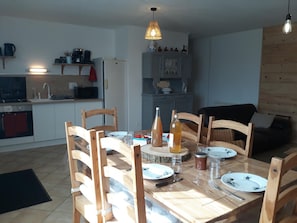 A dining area with a wooden table set for four, surrounded by wooden chairs. A kitchen with a refrigerator, stove, and shelves is visible in the background.