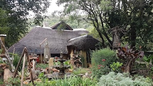 Una casa rústica con techos de paja rodeada de un exuberante jardín con diversas plantas y una pequeña fuente.