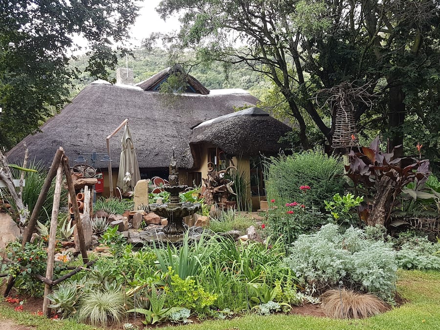 Een rustiek huis met rieten daken, omgeven door een weelderige tuin met diverse planten en een kleine fontein.