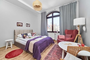 Une chambre moderne avec un lit, un fauteuil rouge, une petite table, une fenêtre avec des rideaux et un luminaire suspendu.