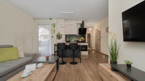 A modern living room with a grey sofa, wooden coffee table, and two black bar stools. A kitchen area with a dining table and chairs is visible in the background.