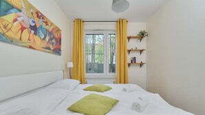 A hotel room with a bed, a window with yellow curtains, a painting on the wall, and a shelf with plants.
