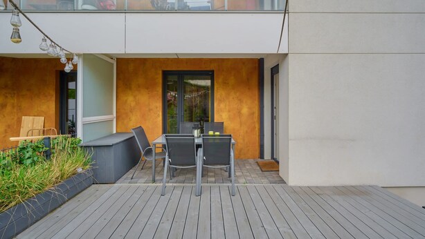A modern outdoor patio with a wooden deck, a table with chairs, and a glass door leading to a balcony.