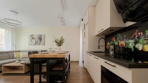 A modern kitchen with a black countertop, white cabinets, and a built-in oven. There is a dining area with a wooden table and chairs, and a sofa in the background.