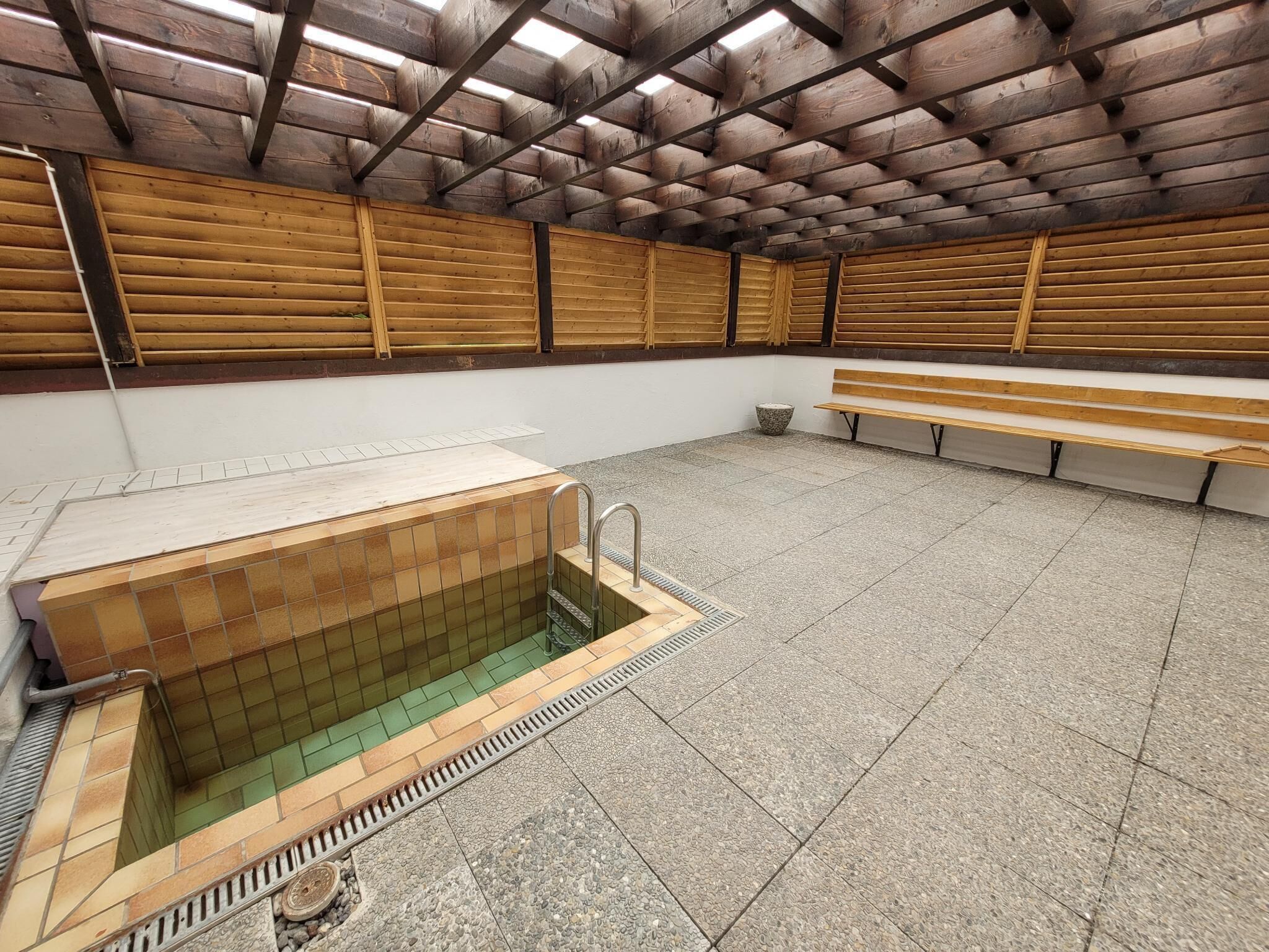 A public bath area with a tiled floor, wooden benches, and a tiled wall.