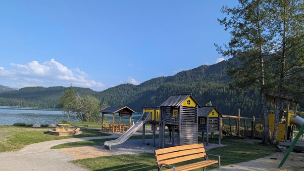 Une aire de jeux avec un toboggan et des cabanes en bois, un banc, et une vue sur un lac et des montagnes.