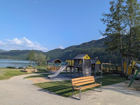 A playground with a slide and wooden playhouses, a bench, and a view of a lake and mountains.. Top Ausgestattete Ferienwohnung mit Schwimmbad und Sauna - 10 Min. zum Weißensee