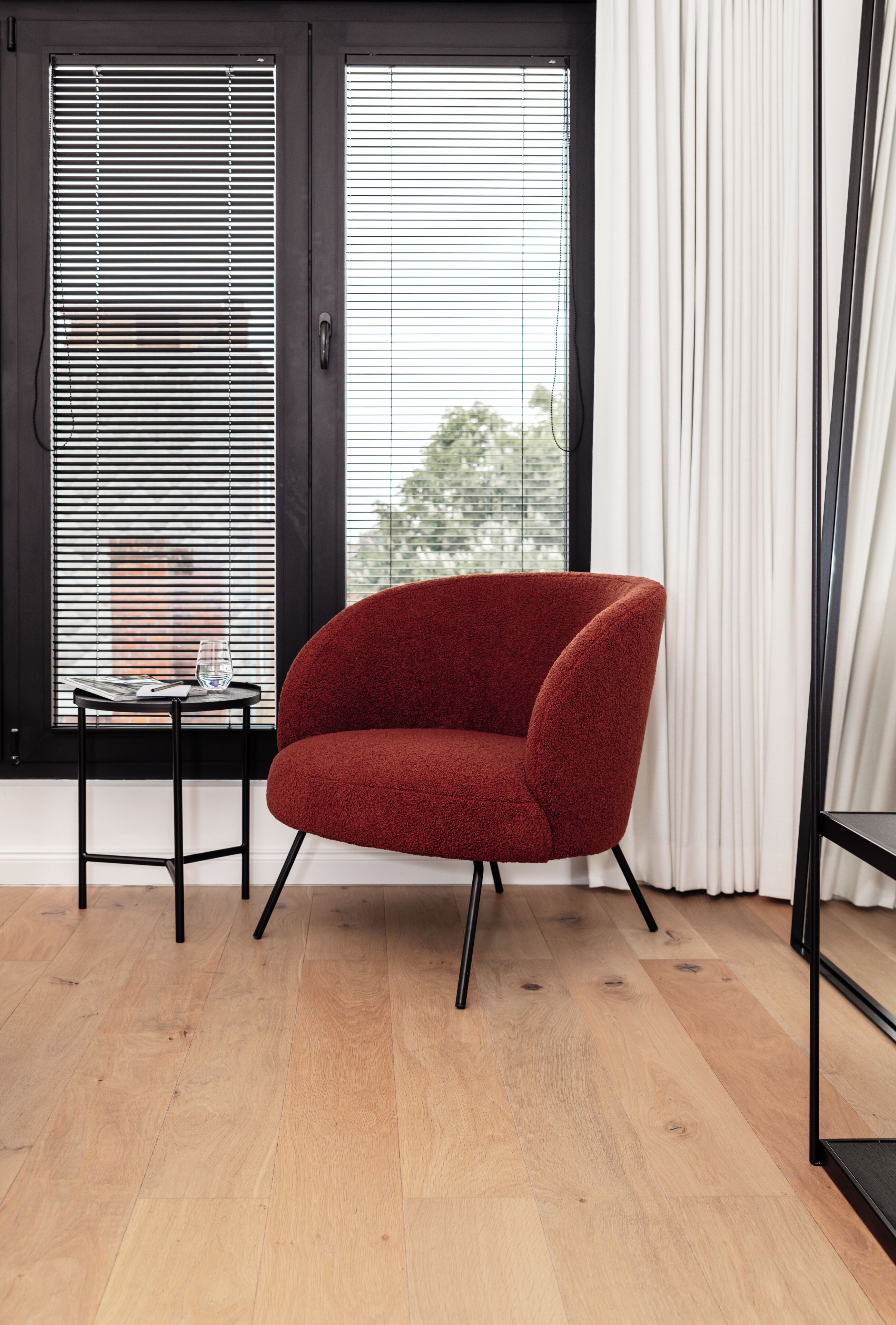 A red armchair in a room with wooden flooring, a small table with a glass, and a window with blinds.