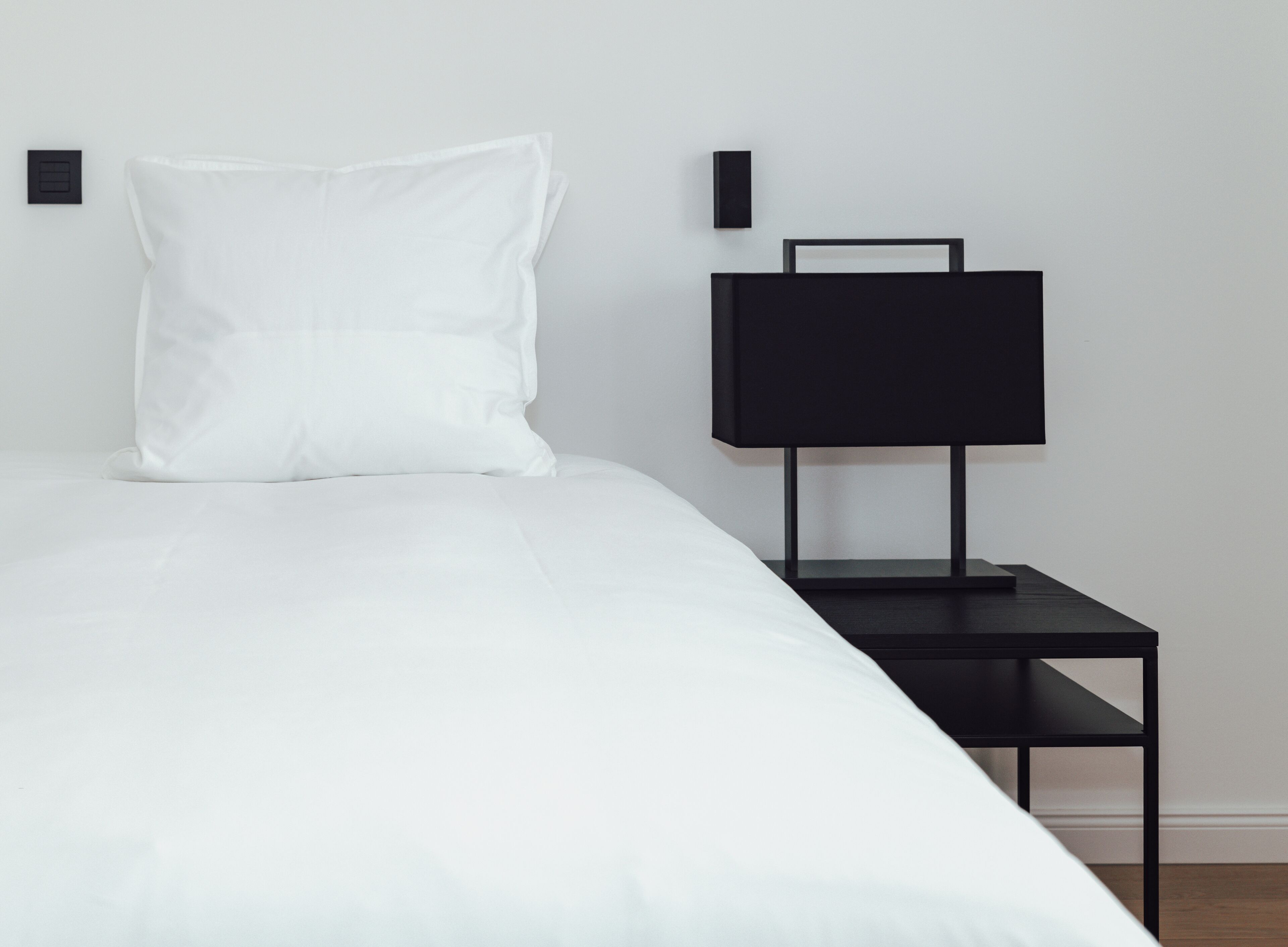 A minimalist bedroom with a white bed, a black nightstand, and a black wall-mounted lamp.