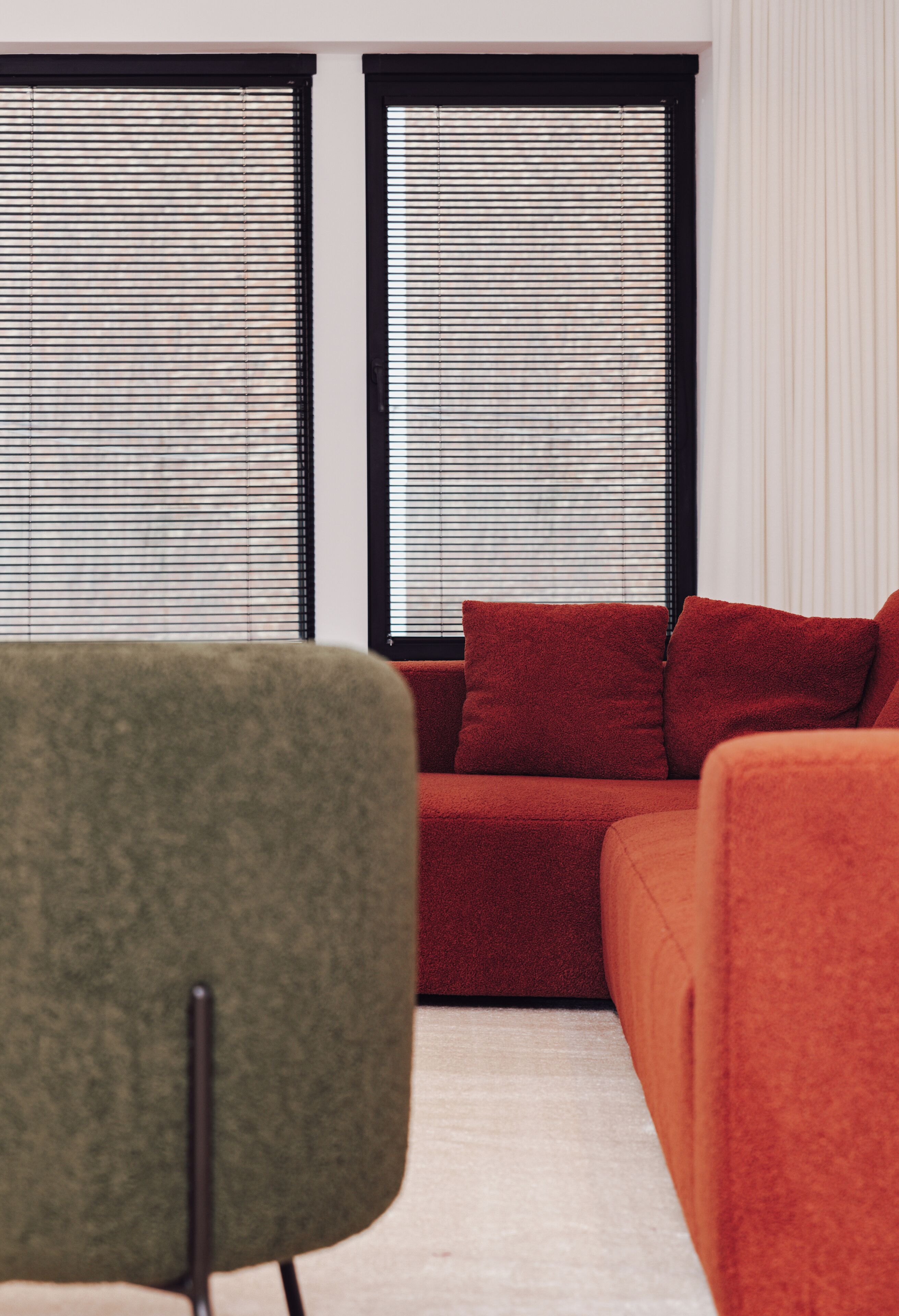 A modern living room with a red sofa, a green armchair, and two windows with blinds.