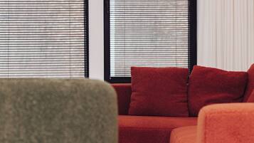A modern living room with a red sofa, a green armchair, and two windows with blinds.