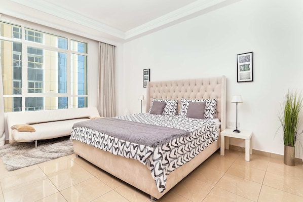 A bedroom with a large bed, a bedside table, a lamp, a potted plant, and a window with curtains.