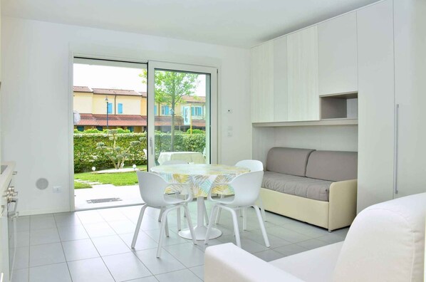 A modern living room with a sofa, a dining table with chairs, and a sliding glass door leading to a garden.