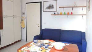 A room with a blue sofa, a dining table with a fruit-themed tablecloth, a white wardrobe, and a door with a towel hanging.
