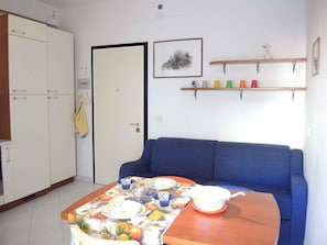 A room with a blue sofa, a dining table with a fruit-themed tablecloth, a white wardrobe, and a door with a towel hanging.
