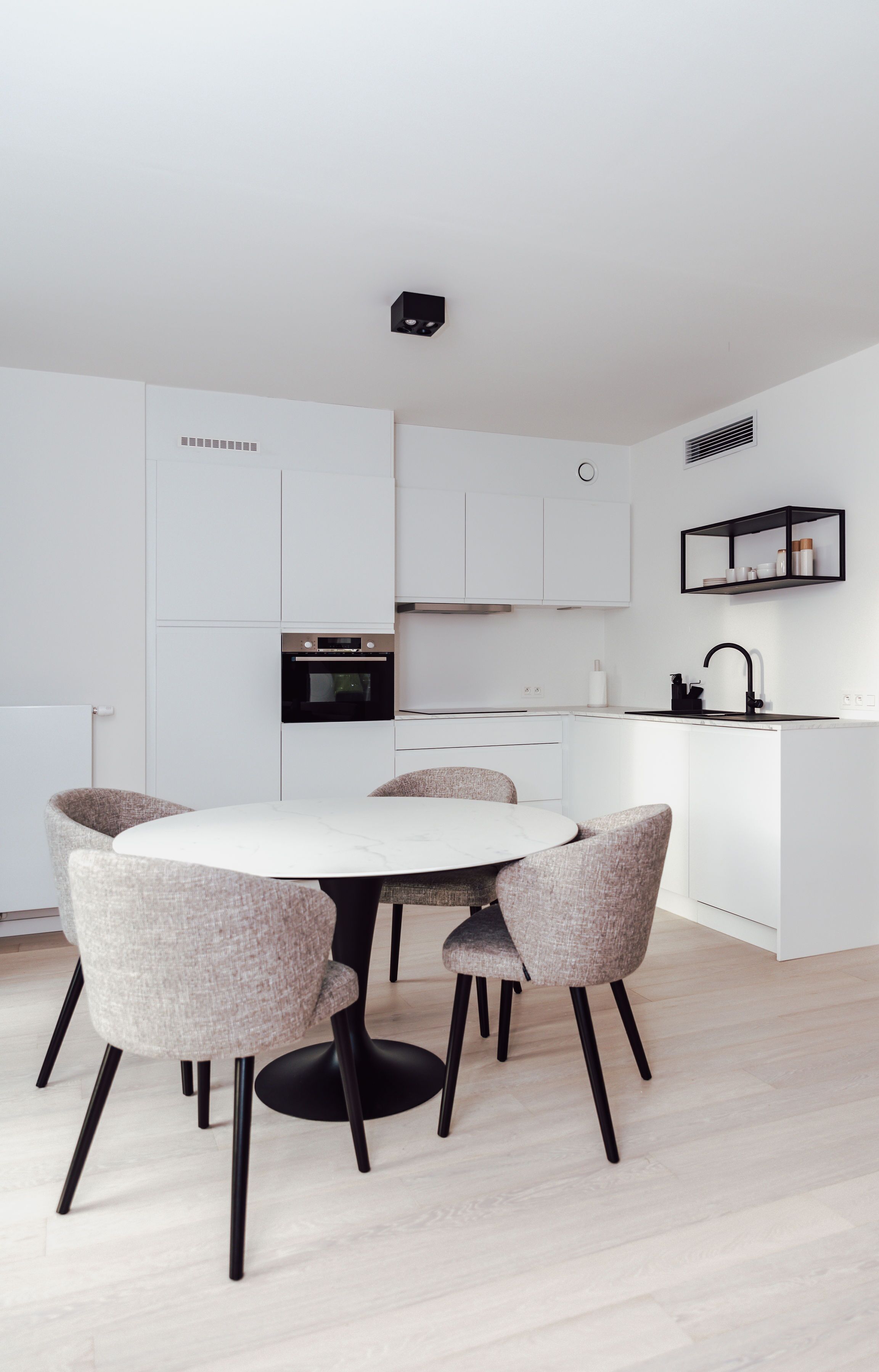 A modern kitchen with white cabinetry, a round dining table with four chairs, and a black countertop with a sink and a shelf.