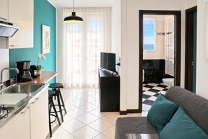 A modern kitchen with a coffee maker, a dining area with a table and chairs, a sofa with teal cushions, and a view of the sea through a window. - Leone sea view apartments on the Rimini seafront (Rimini)