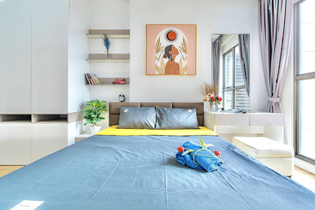 Ein modernes Schlafzimmer mit einem großen Bett, einem Nachttisch, einer kleinen Topfpflanze, einem an der Wand befestigten Regal mit Büchern und einem Fenster mit Vorhängen.